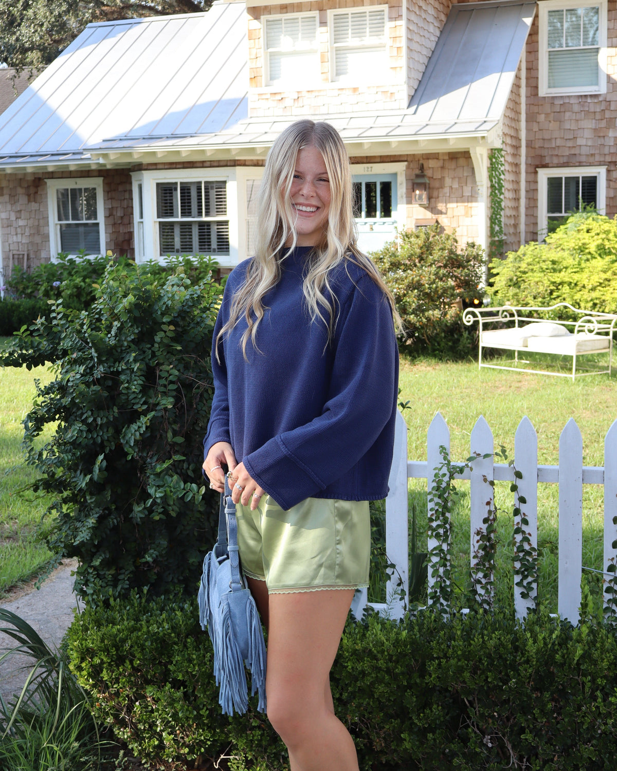 A woman stands on a sidewalk in front of a suburban house, wearing a navy blue sweatshirt, shiny olive green shorts, and holding a light blue tassel bag.